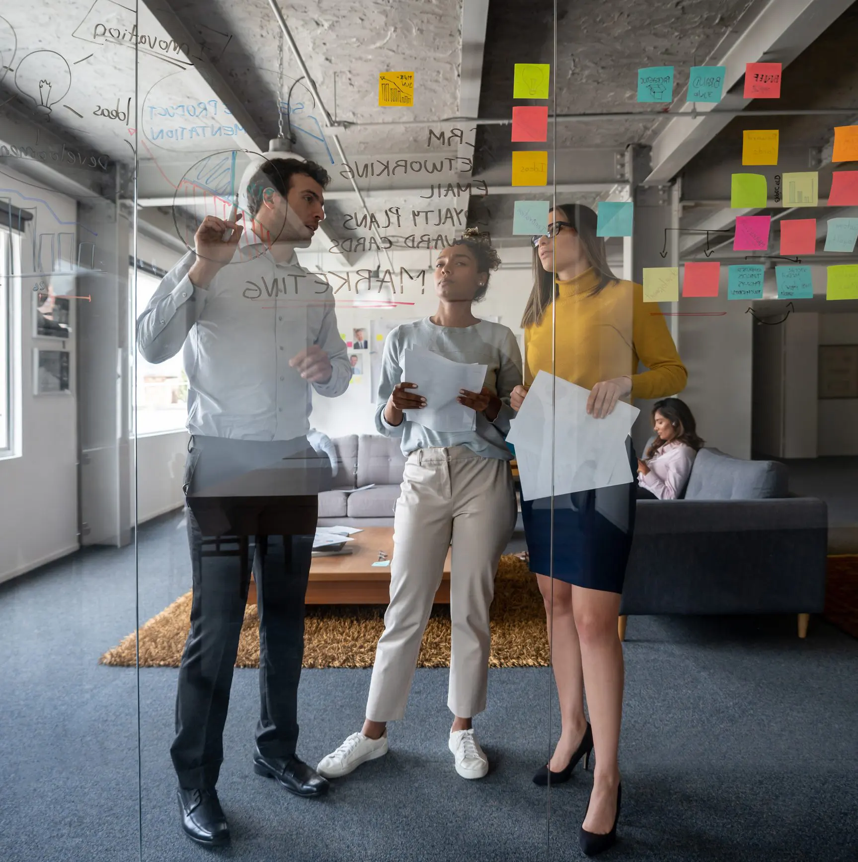 Three colleagues brainstorming with colorful sticky notes on a glass wall in a modern office.