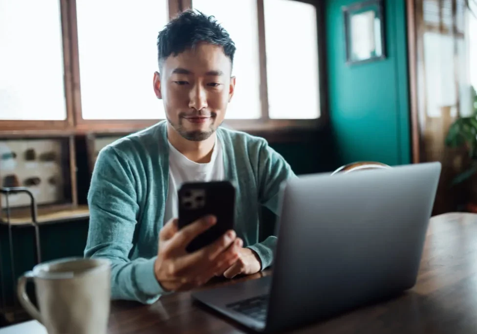 Man sitting at desk, looking at smartphone with laptop nearby.